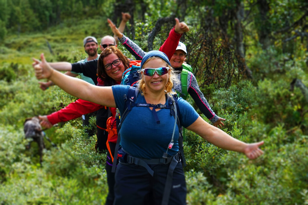 Smildende og glade turgåere på rekke langs en skogsti som strekker armene ut. Omgitt av grønn skog.