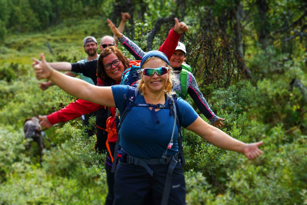 Smildende og glade turgåere på rekke langs en skogsti som strekker armene ut. Omgitt av grønn skog.
