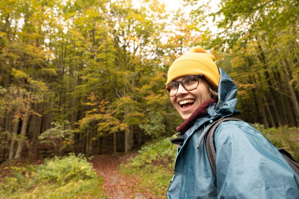 Young adult woman in autumn forest wearing a yellow beanie and blue jacket, smiling joyfully during a hike.