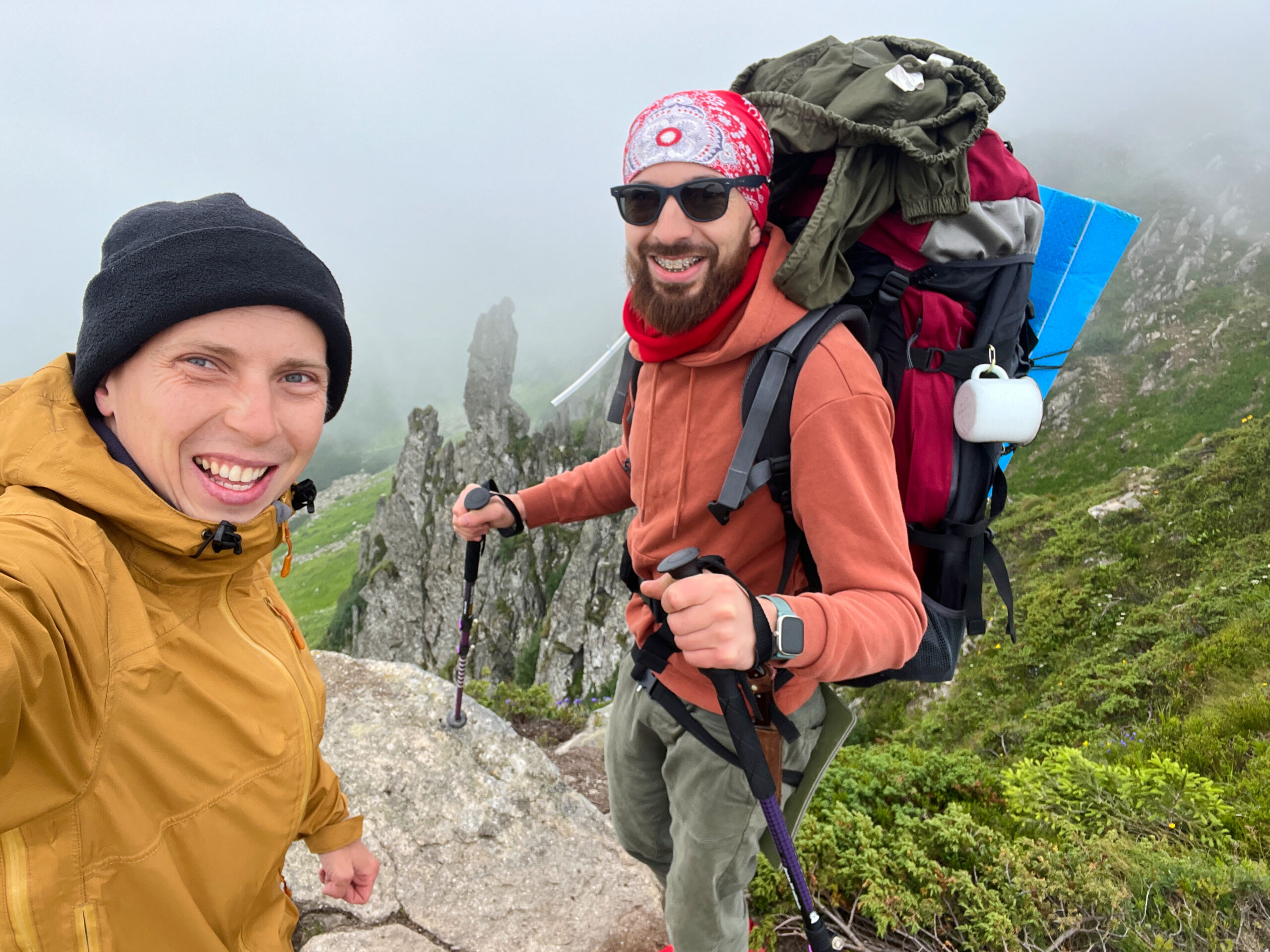 Two smiling hikers take selfie on misty mountain trail, equipped with large backpacks and trekking poles, with rugged cliffs visible in background.