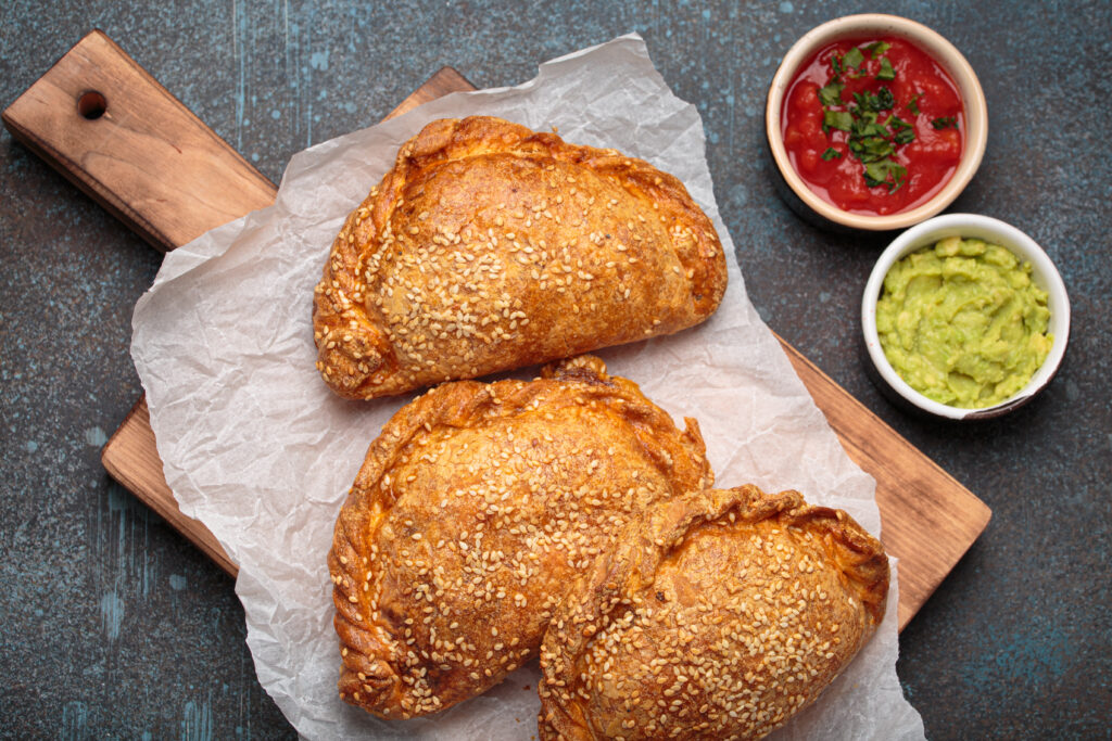 Three freshly baked empanadas with sesame seeds on a wooden board, served with salsa and guacamole. Traditional dish of Spain and Latin America