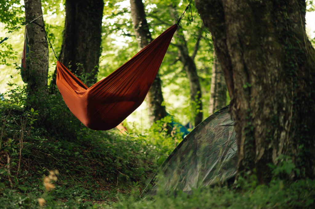 A person is sleeping and resting in a hammock in a forrest next to a tent. Nature lover is enjoying summer day in woods and taking a break from wild night on an open air festival.