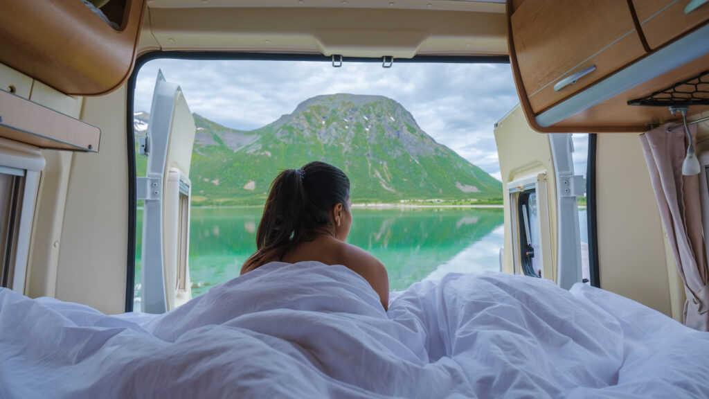 A woman lies in bed in a camper van, gazing out the window at a stunning mountain view and the turquoise water of a fjord in Norway. Lofoten Norway