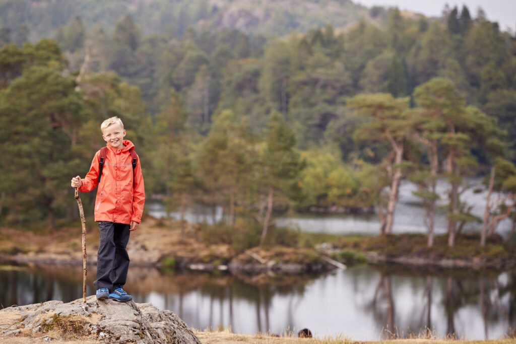 A boy standing on a rock by a lake holding a stick, smiling to camera, Lake District, UK